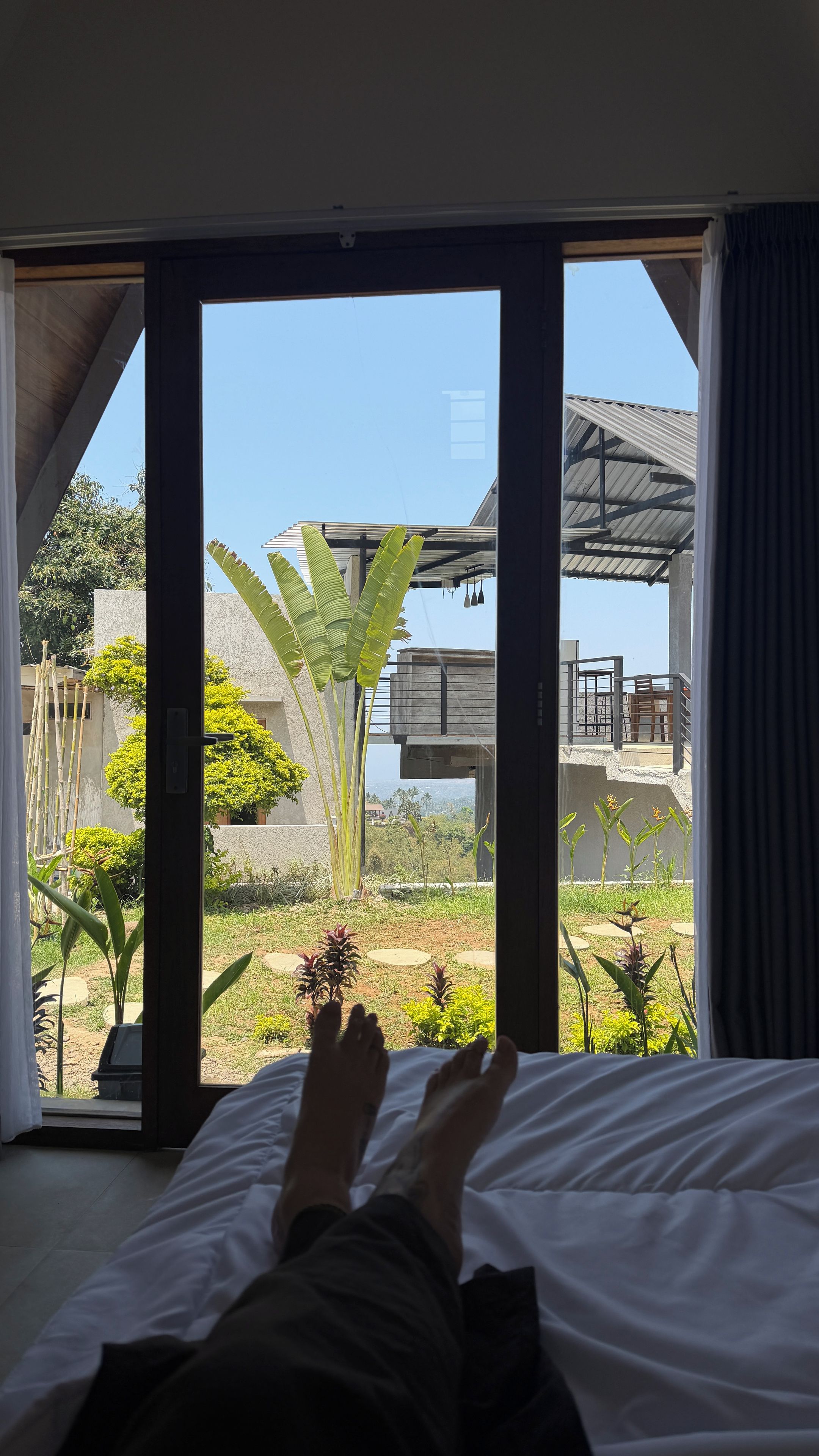 A room with a bed, a view through a glass door to an outdoor area with plants and a building.