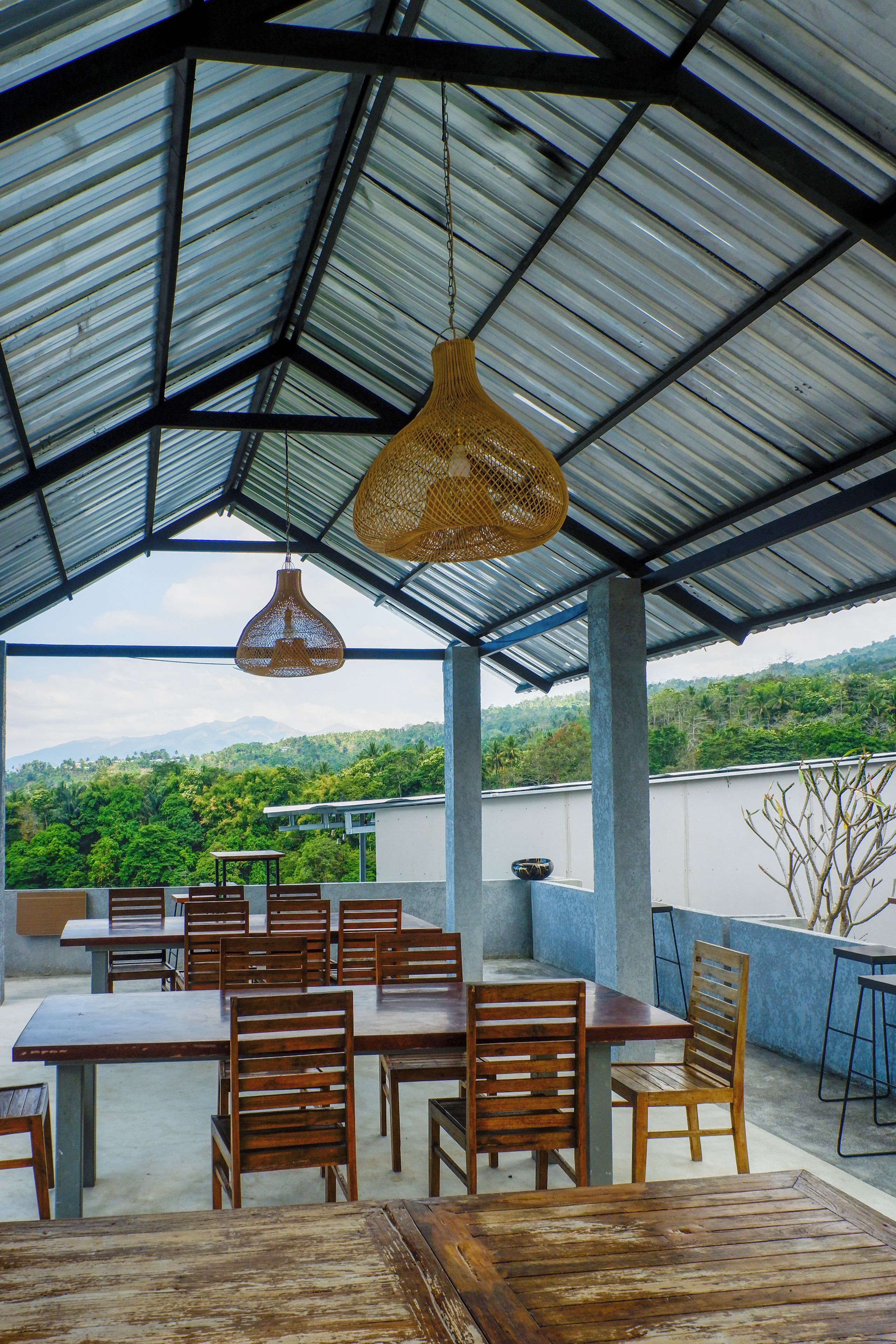 An outdoor dining area with tables and chairs, a view of a green landscape, and a metal roof structure.