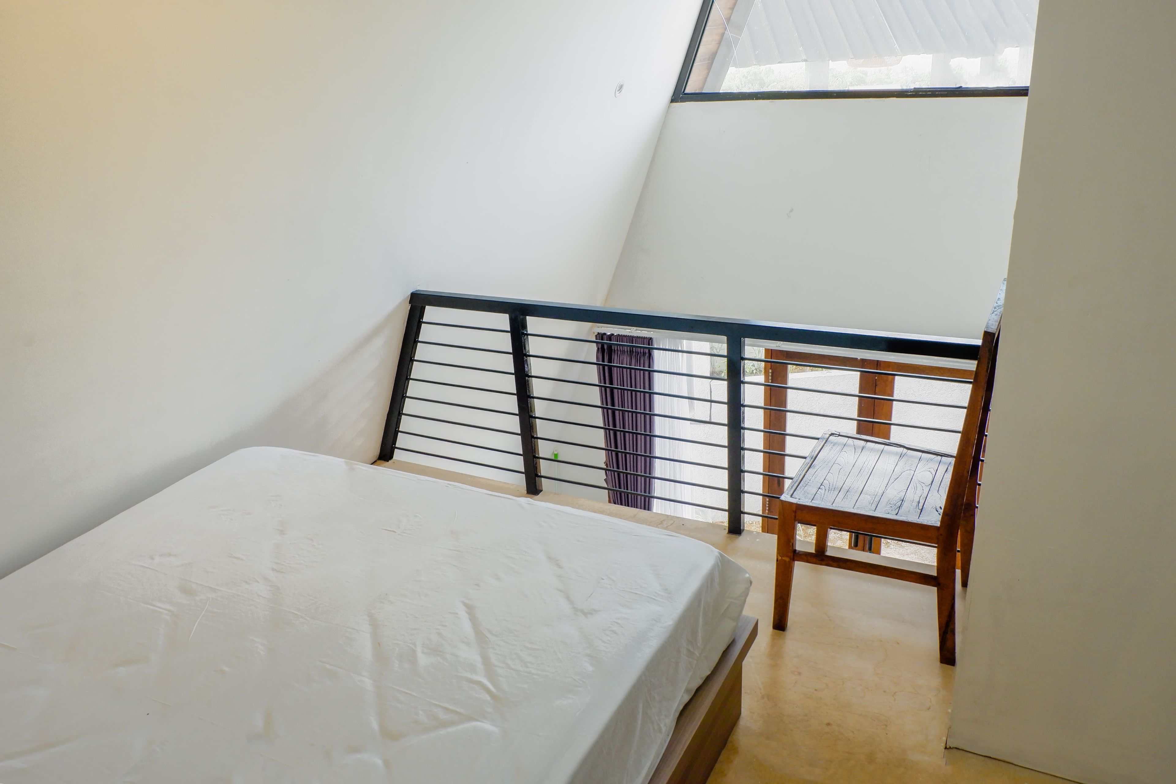 A small, single-bed room with a wooden chair and a window with a metal railing.