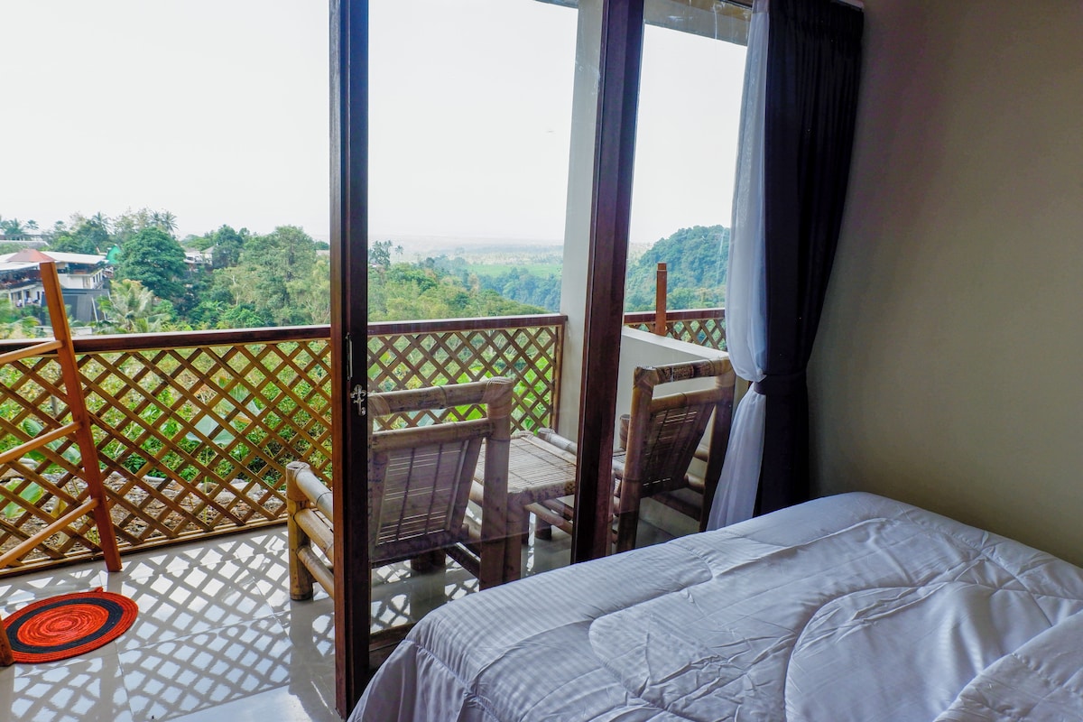 A room with a bed, a table, and a chair. There is a view of greenery and buildings outside the window.