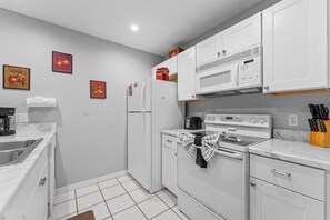 Een kleine keuken met witte kastjes, een koelkast, een magnetron en een fornuis. Twee ingelijste foto's hangen aan de muur en er staat een koffieapparaat op het aanrecht.