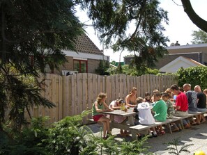 An outdoor gathering with people seated at wooden tables, surrounded by a wooden fence and greenery. - Holiday Apartment With 2 Bedrooms (Renesse)