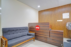Un área de recepción con un mostrador de madera, un sofá gris y un ventilador fijado a la pared.
