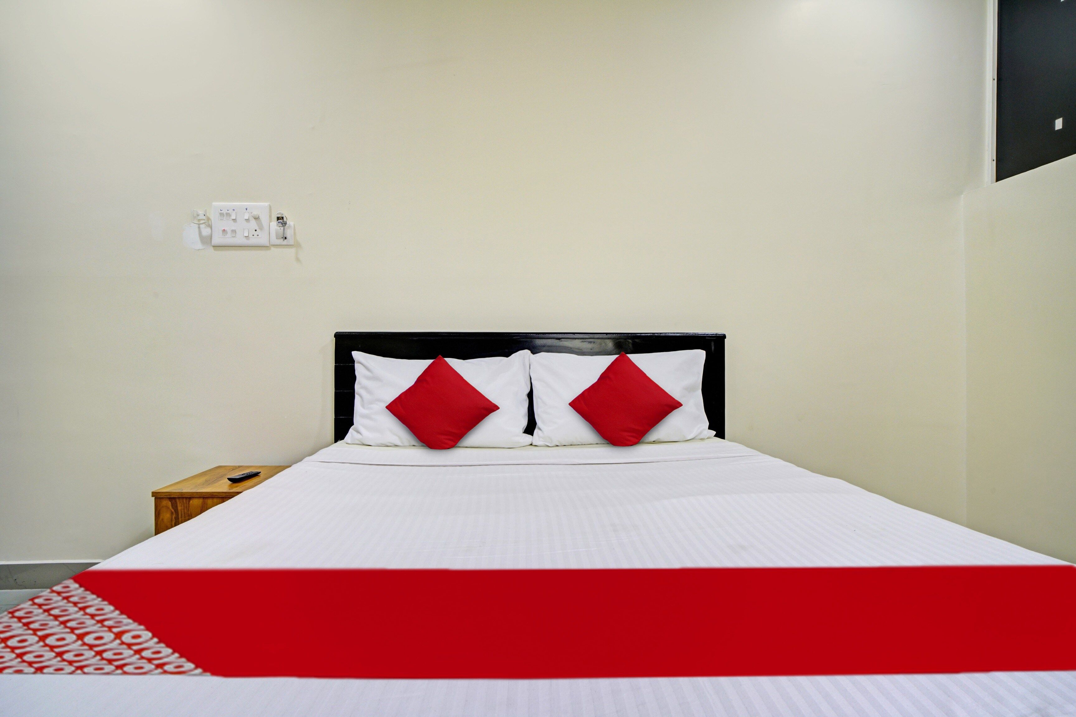 Una cama con colchón blanco y dos almohadas rojas, una mesita de noche de madera con un control remoto y una pared blanca con un enchufe.