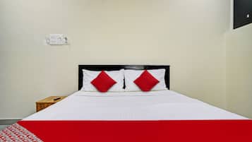 Una cama con colchón blanco y dos almohadas rojas, una mesita de noche de madera con un control remoto y una pared blanca con un enchufe.