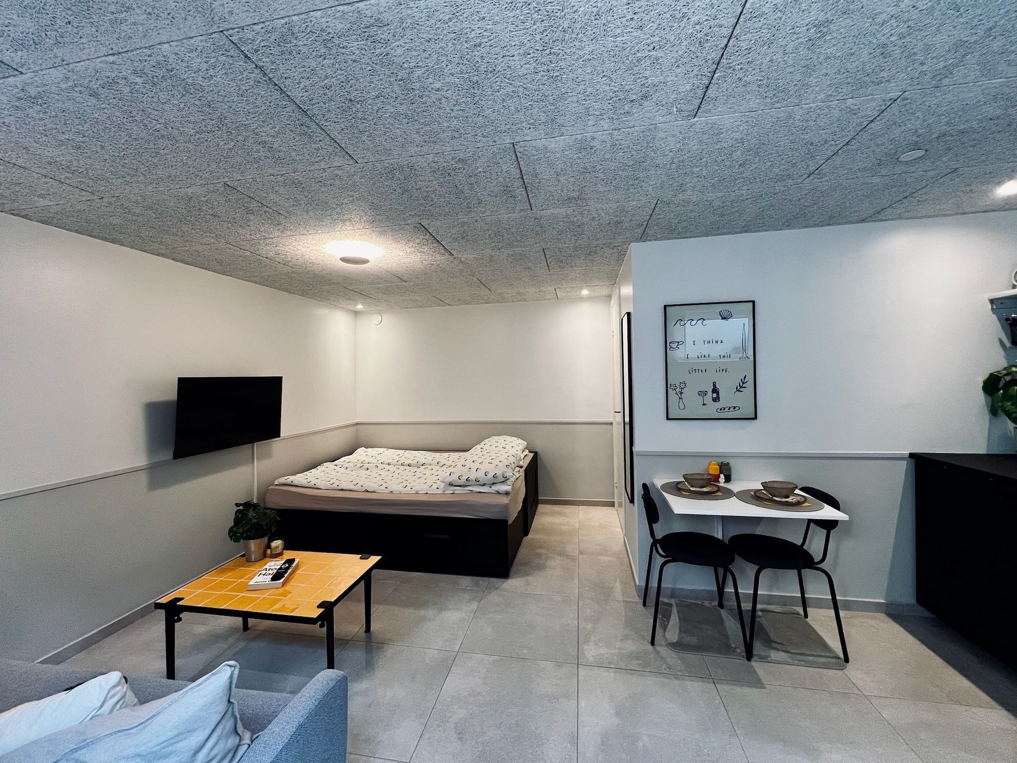 Een knus woongedeelte met een bed, een klein tafeltje, een aan de muur bevestigde televisie en een eethoek met stoelen.