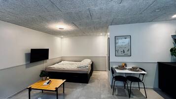 Een knus woongedeelte met een bed, een klein tafeltje, een aan de muur bevestigde televisie en een eethoek met stoelen.