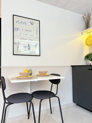 A modern kitchen with a white table, two black chairs, a framed artwork, and a yellow pot on the wall. - New Cozy Basement Studio Apartment! (Rødovre)