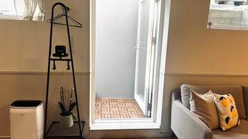 Ein Zimmer mit einer Couch, einem Heizkörper, einem kleinen Tisch, einer hängenden Pflanze und einem Fenster mit Blick auf eine Ziegelwand.
