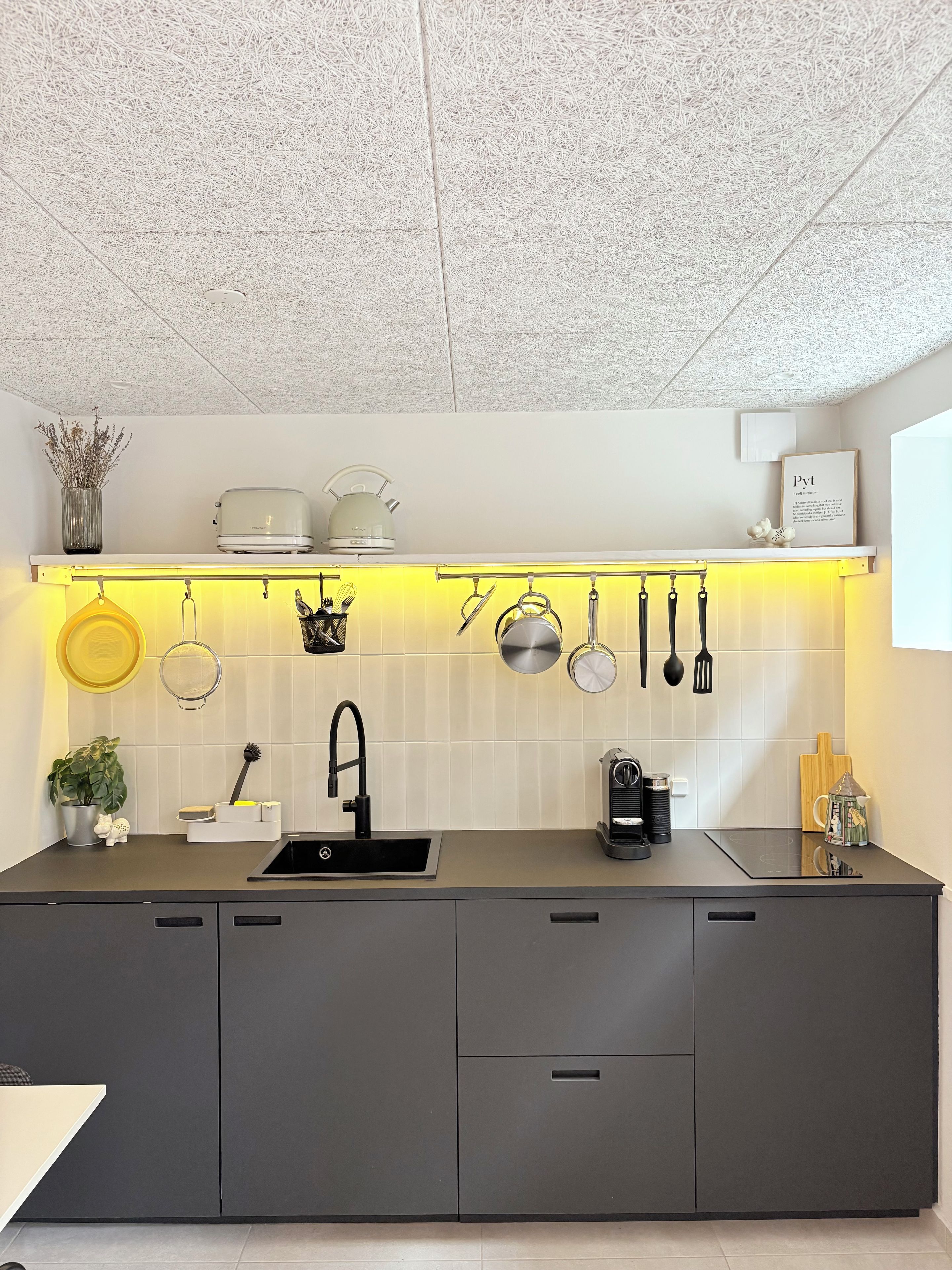 Eine moderne Küche mit schwarzer Arbeitsplatte, weiß gefliestem Spritzschutz und einem gelb beleuchteten Regal über dem Spülbecken.