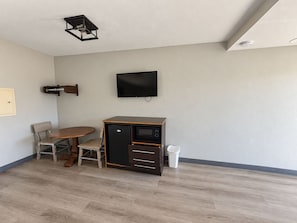 Ein kompakter Raum mit einem wandmontierten Flachbildfernseher, einer kleinen Küchenzeile mit Kühlschrank und Mikrowelle, einem Holztisch mit Stühlen und einer weißen Mülltonne.