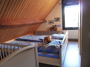 A small, cozy room with a sloped ceiling, a single bed with a blue and yellow patterned bedspread, a teddy bear on the bed, and a window with a view of the outside. - Holiday Home With 1 Bedroom (Oostkapelle)