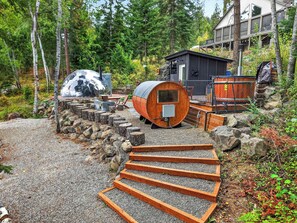 Un espacio exterior rústico con un barril de madera, una cabaña pequeña y un sendero de piedra que lleva a una zona de estar.
