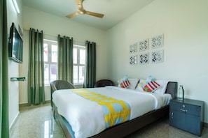 Quarto de hotel com cama, escrivaninha, cadeira, televisão e ventoinha no teto.