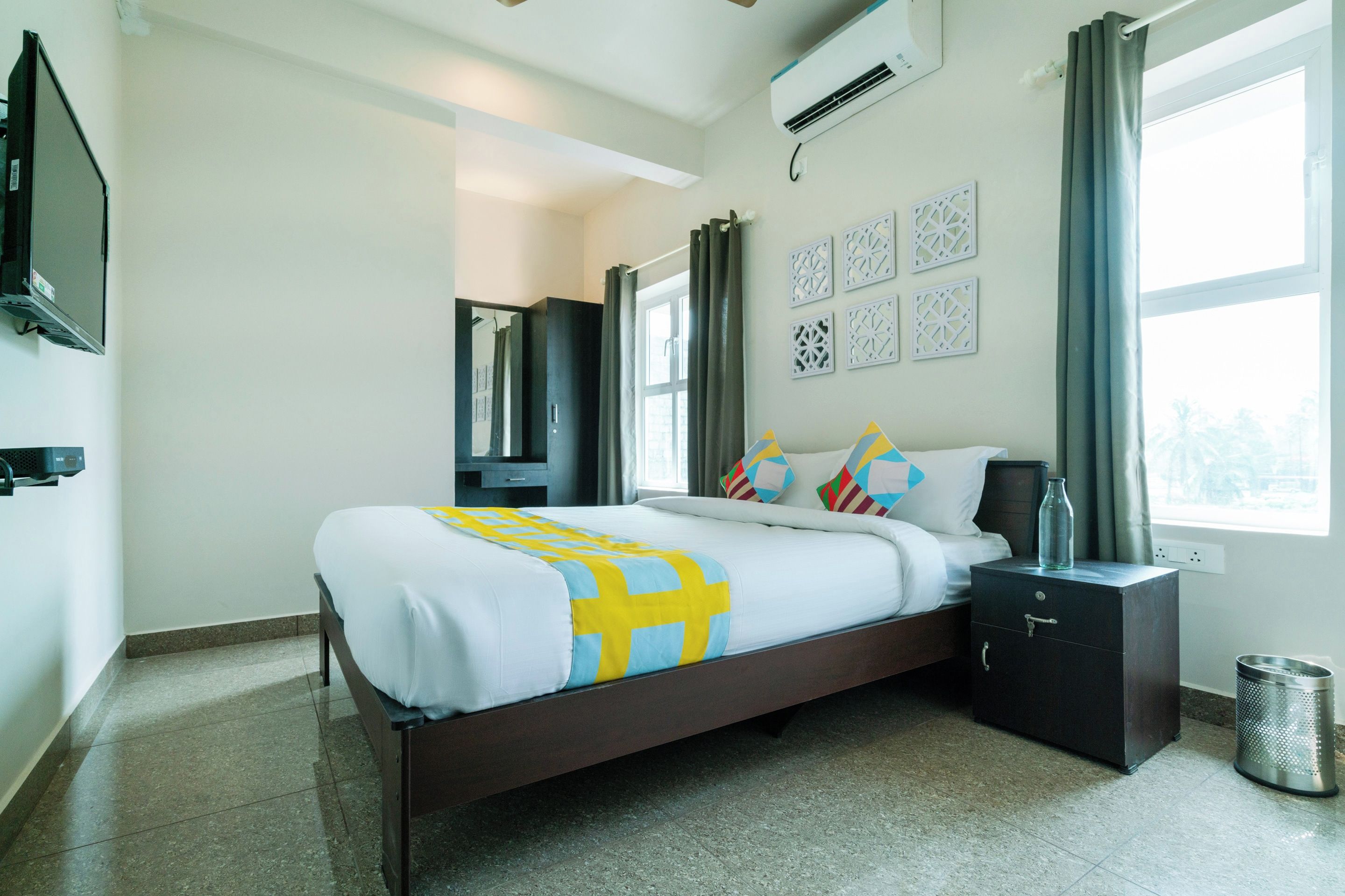 Een hotelkamer met een bed, nachtkastje, televisie, raam met gordijnen en een wandairconditioner.