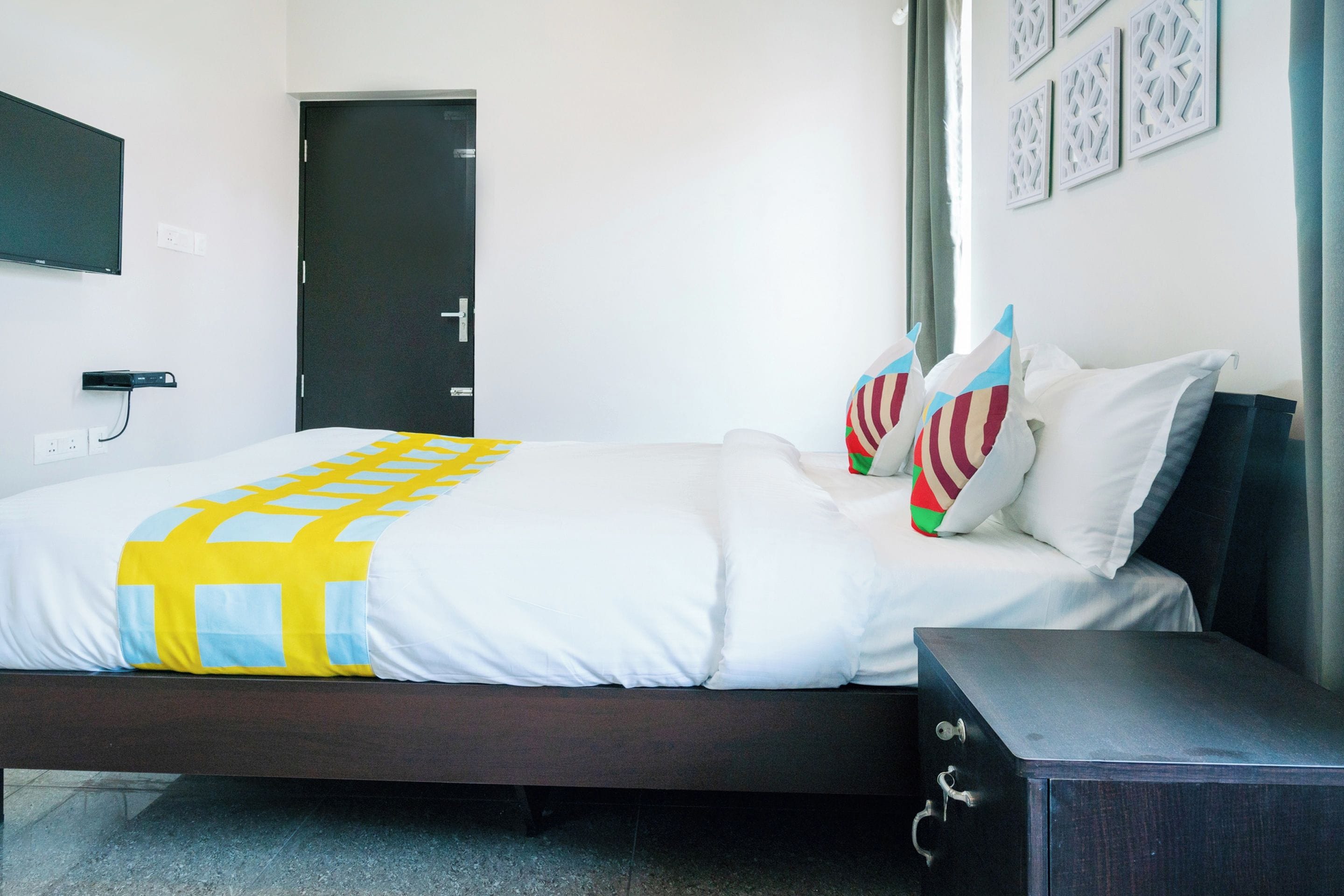 A hotel room with a bed, a bedside table, a TV mounted on the wall, and a door.
