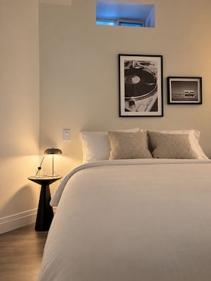 Ein Schlafzimmer mit einem Bett, Kissen, einem Nachttisch mit Lampe und zwei gerahmten Bildern an der Wand.
