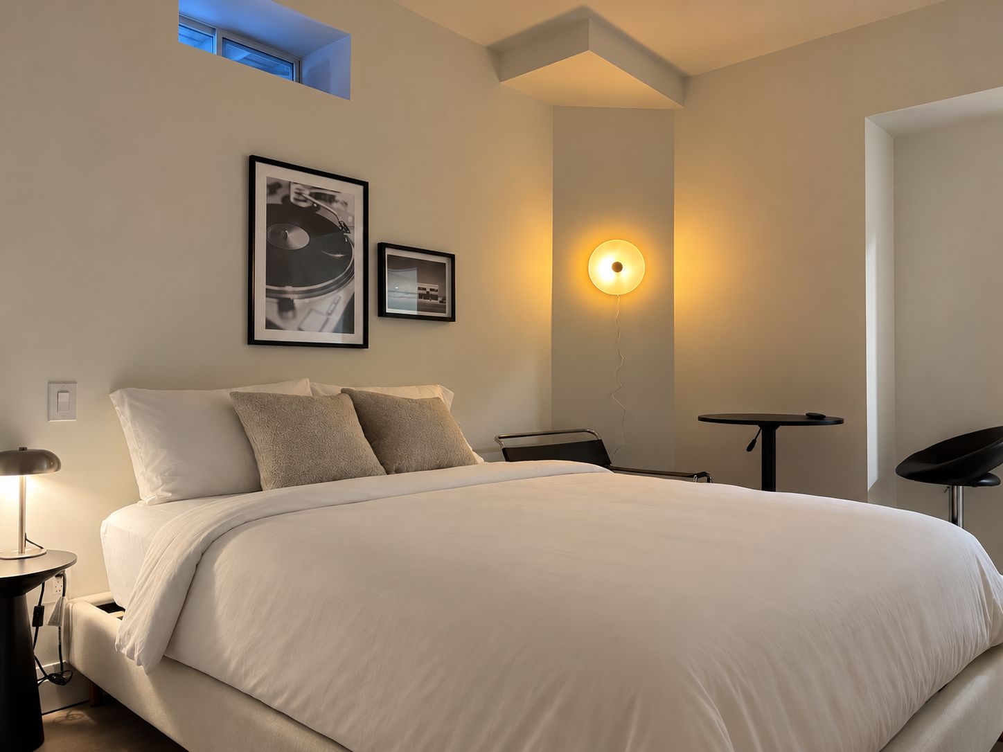A modern bedroom with a large bed, a bedside table, a desk, and a chair. There are two framed pictures on the wall.