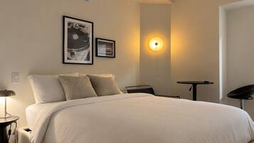 A modern bedroom with a large bed, a bedside table, a desk, and a chair. There are two framed pictures on the wall.