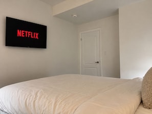 Un dormitori amb un llit, una porta i una televisió penjada a la paret que mostra 'NETFLIX'.