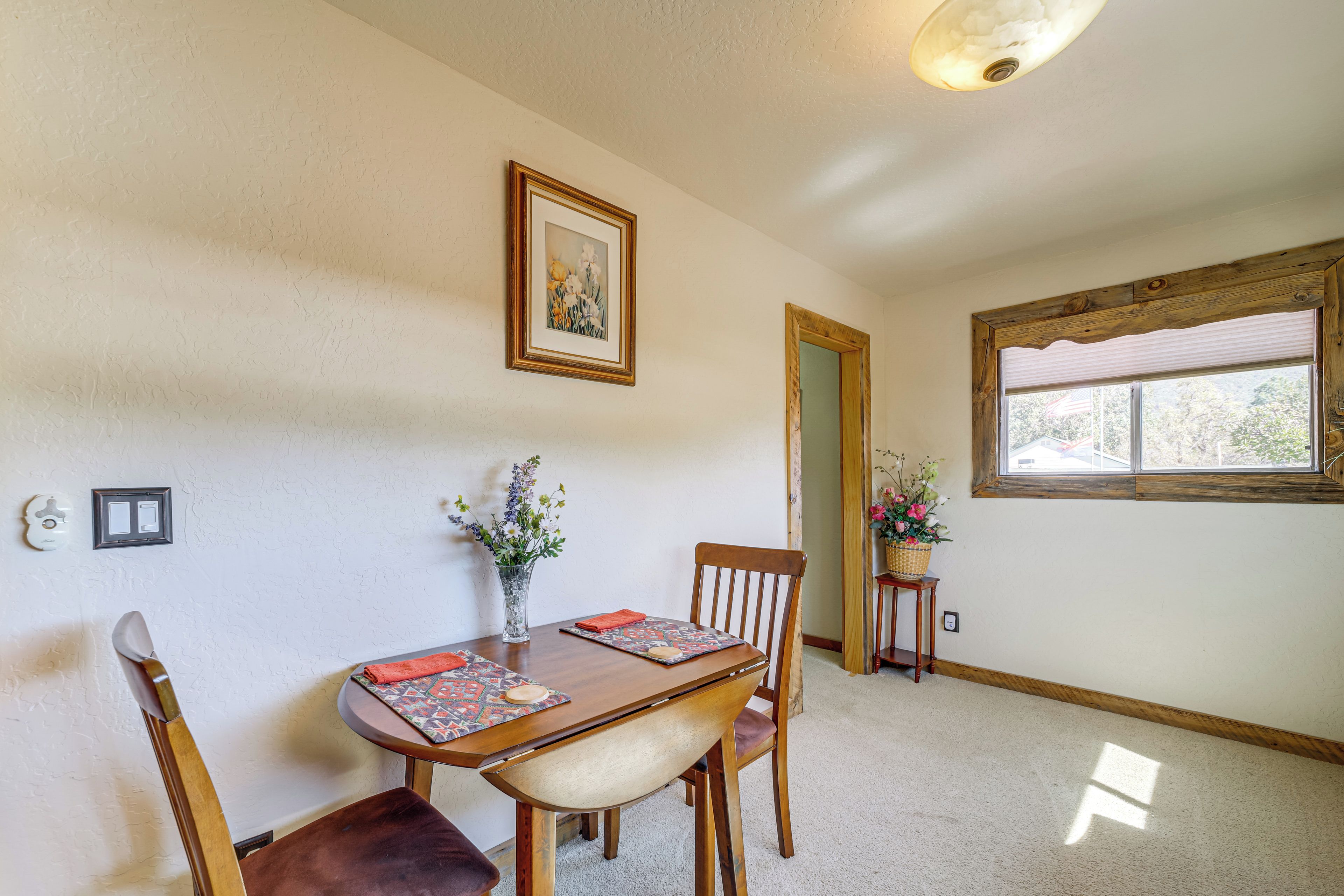 A dining area with a wooden table, chairs, a vase with flowers, a framed picture on the wall, and a window with blinds.