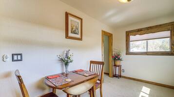 A dining area with a wooden table, chairs, a vase with flowers, a framed picture on the wall, and a window with blinds.