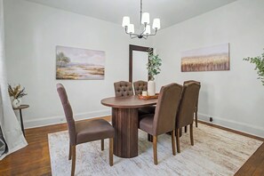 Un comedor con una mesa de madera redonda, seis sillas tapizadas, una lámpara de araña y dos cuadros de paisajes en las paredes.