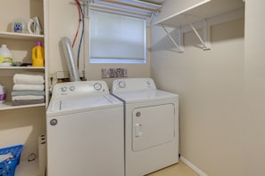 Une buanderie équipée de deux machines à laver à chargement frontal blanches, d’une étagère contenant des produits de lessive, d’une fenêtre avec des stores et d’un étendoir à linge.