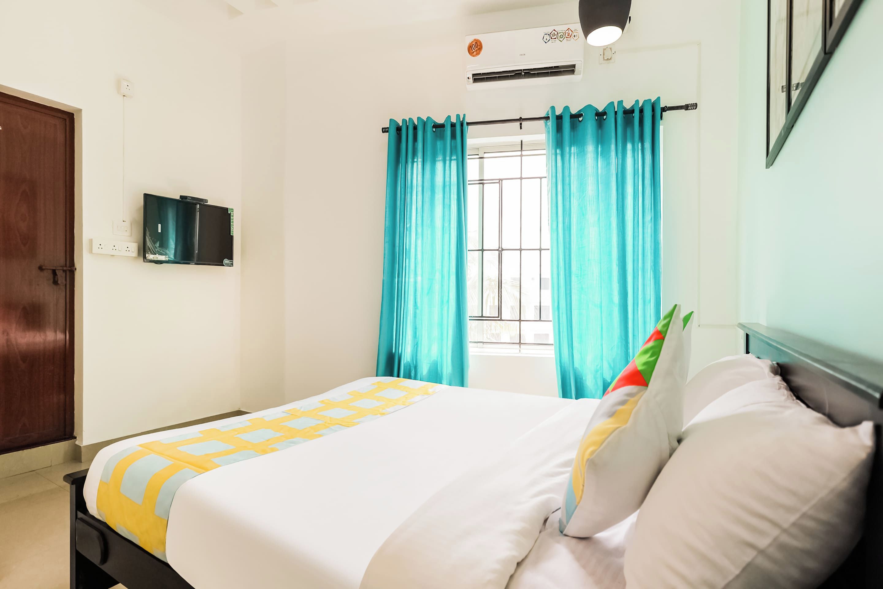 Habitación de hotel con cama, ventana con cortinas, aire acondicionado y televisor montado en la pared.
