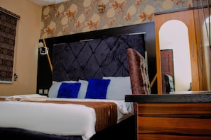 Une chambre d’hôtel avec un lit, une commode en bois, une tête de lit capitonnée de couleur sombre et un papier peint à motifs.