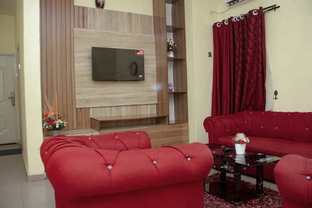 A living room with a red sofa set, a flat-screen TV mounted on a wooden wall, and a red curtain.