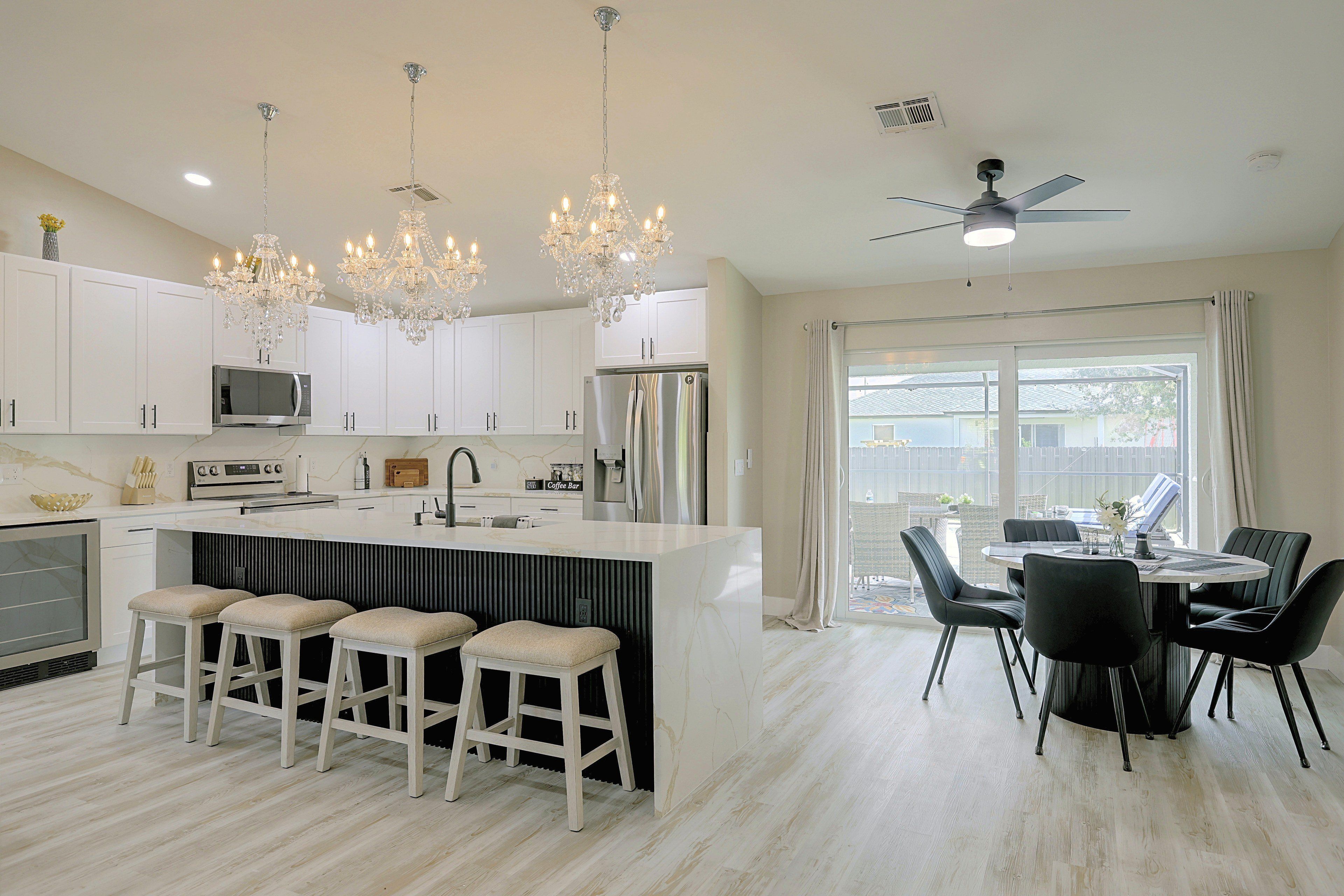 A modern kitchen with white cabinets, a central island with stools, a dining area with a table and chairs, and a ceiling fan.