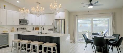 A modern kitchen with white cabinets, a central island with stools, a dining area with a table and chairs, and a ceiling fan.