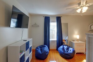 A room with a flat-screen TV mounted on the wall, a ceiling fan, a window with curtains, a bedside table with a lamp, and a wicker chair. - Beach Escape 1 Mi to Okaloosa Island & Dtwn (Fort Walton Beach)