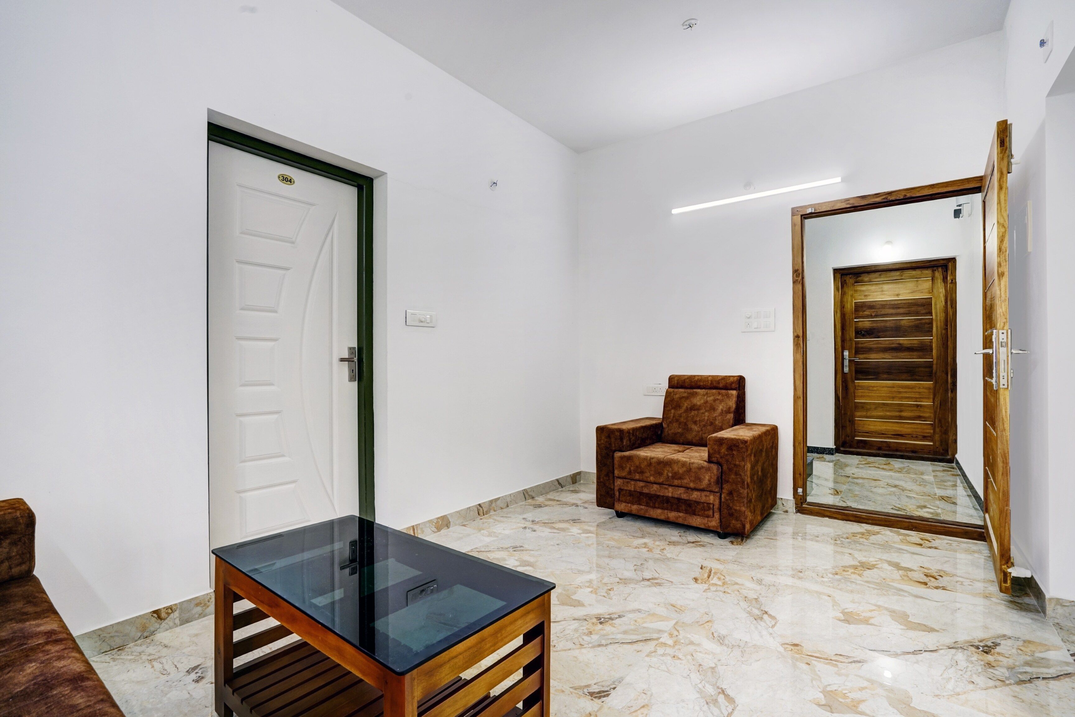 A room with a white door, a brown armchair, a glass-top table, and a wooden-framed mirror on the wall.