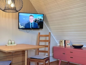 A room with a television mounted on the wall, a wooden chair, a wooden table, a pink cabinet, and a pendant light. - defaultName (Glowe)