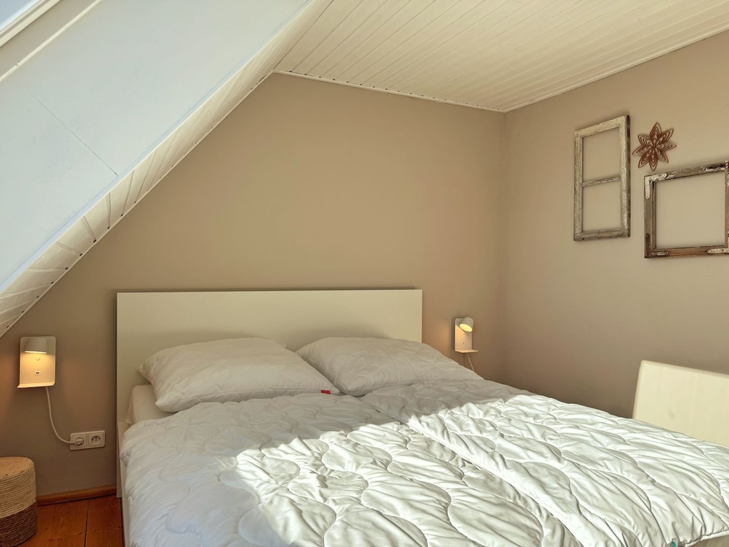 A bedroom with a sloped ceiling, a bed with white bedding, two wall-mounted lamps, and two picture frames on the wall.