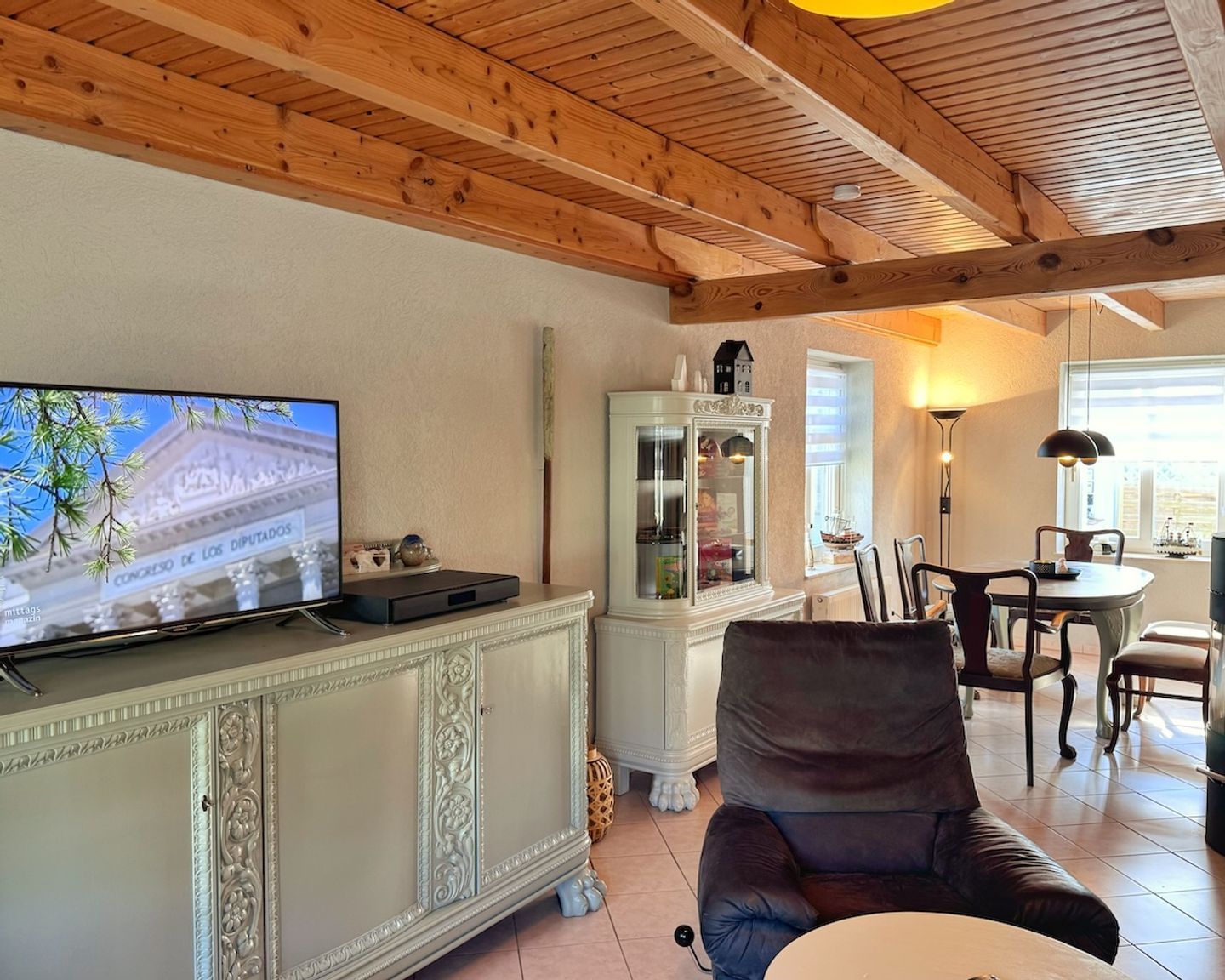 A living room with a television displaying a building, a cabinet with glass doors, a dining area with chairs, and a ceiling with wooden beams.