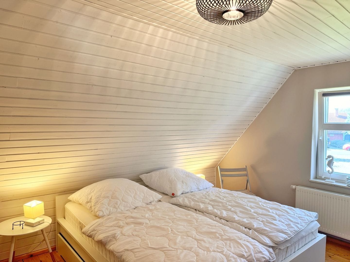 Une chambre avec un plafond en pente, un lit avec une literie blanche, une table de chevet avec une lampe et une fenêtre donnant sur l’extérieur.