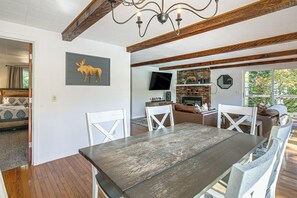 A dining area with a wooden table and white chairs, a television mounted on the wall, and a brick fireplace. - McKenzie River Home w/ Waterfront Fire Pit (Vida)