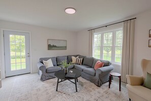 A living room with a grey sectional sofa, a coffee table, and a small side table. There is a door with a view of greenery outside, a window with curtains, and a painting on the wall. - 50-Acre Hilltop Home: Upstate Getaway (Mooers)