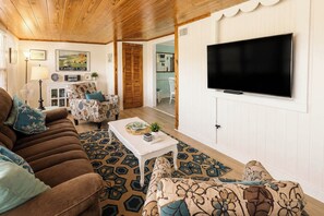 Een woonkamer met een bruine bank, gestreepte fauteuils, een witte salontafel en een flatscreen televisie aan de muur.
