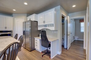 A modern kitchen with white cabinetry, a stainless steel refrigerator, a dining table with chairs, and a built-in office space with a chair. - Extended Mtn-View Stay w/ Hot Tub Near Skiing (Mountain Green)