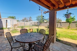A covered patio with a round table and chairs, surrounded by a metal fence, with a view of a backyard and a building in the background. - Studio 3 Mi to Tucson Airport! Bring Your Pups (Tucson)