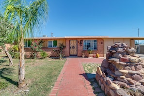 A single-story house with a brick facade, a front door with a decorative metal design, a small front yard with a palm tree, and a stone wall to the right. - Studio 3 Mi to Tucson Airport! Bring Your Pups (Tucson)