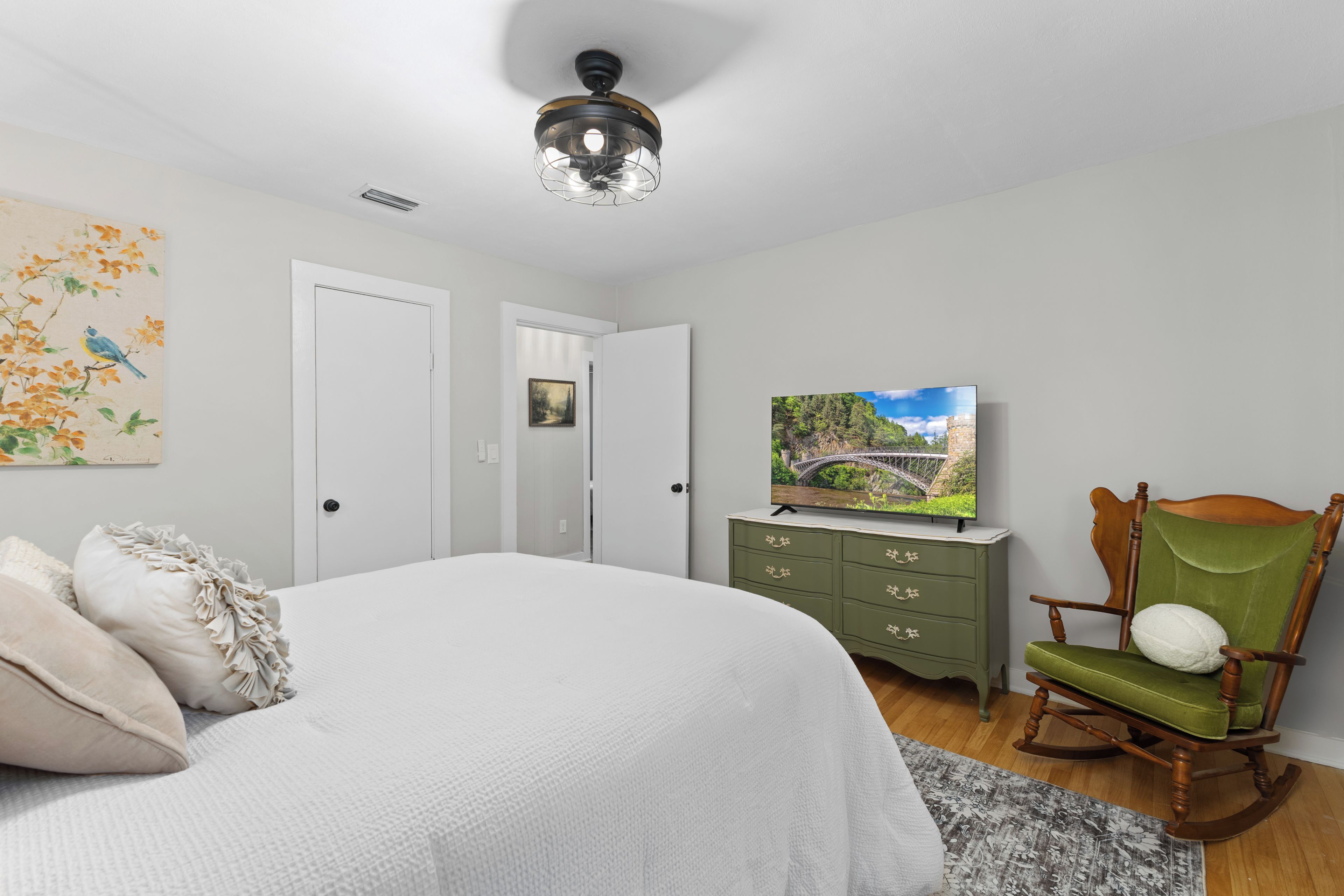 A bedroom with a bed, a television on a dresser, a rocking chair, and a painting on the wall.