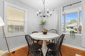 A dining area with a white table, four chairs, a chandelier, a window with blinds, and a view of the outside. - Cute, Cozy & Near Downtown! Updated Home w/ Porch (Live Oak)