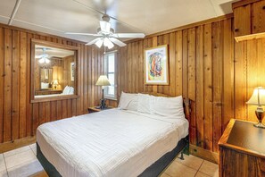 Une pièce aux murs recouverts de lambris en bois, un lit avec une literie blanche, un ventilateur de plafond, une table de chevet avec une lampe et une œuvre d’art encadrée accrochée au mur.