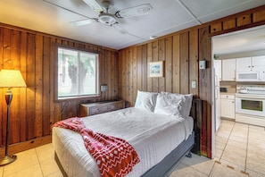 A bedroom with a bed, a nightstand, a lamp, a window with blinds, wooden walls, a ceiling fan, and an adjoining kitchen with appliances. - License to Chill: Lake Access, 14 Mi to Disney (Clermont)