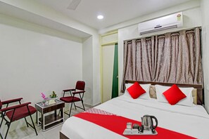 A hotel room with a bed, two red chairs, a small table with a vase of flowers, and an air conditioning unit on the wall.